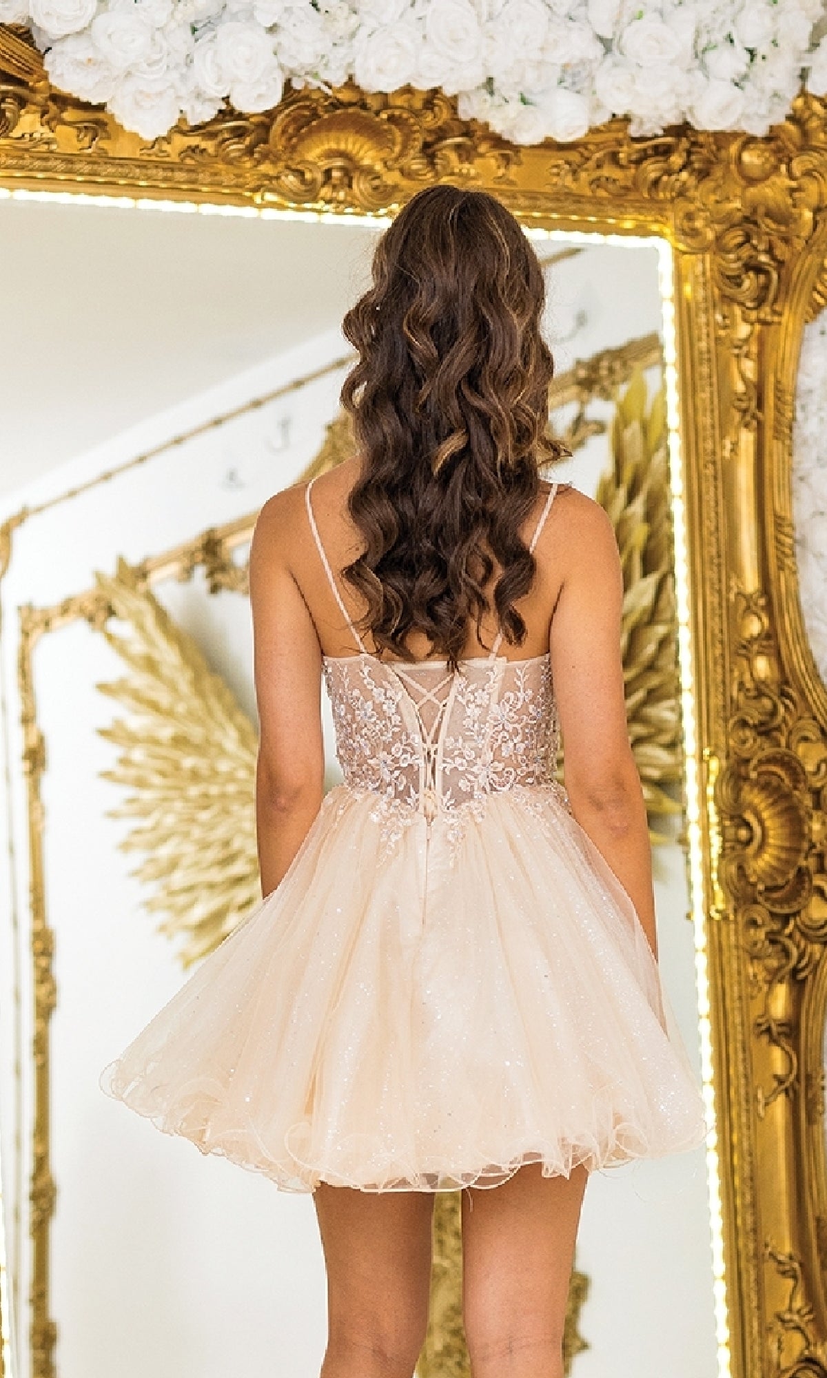 A woman with long, wavy hair wearing the Short Glitter Prom Dress: Dancing Queen 3411 stands facing a large ornate mirror adorned with gold wings and floral decorations.
